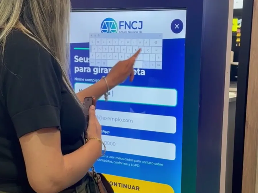 Participante tocando a tela do totem do jogo da roleta no estande do FNCJ